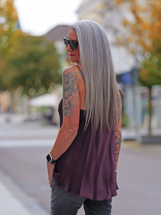 Person mit langen grauen Haaren trägt das OFF#DLY Top Button bordeaux, ein ärmelloses, fließendes Viskose-Oberteil mit Knopfleiste, kombiniert mit dunklen Jeans und einer Smartwatch.