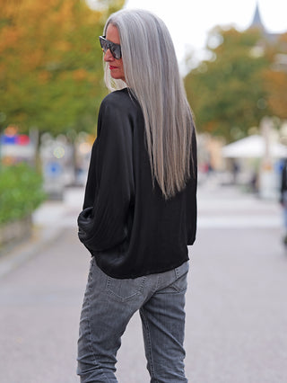 Person trägt das OFF#DLY LS Shirt Valentina black mit fließendem Stoff und weiten Ärmeln. Sie steht auf einer baumgesäumten Straße, leicht abgewandt, Hände in den Hosentaschen, im urbanen Ambiente.