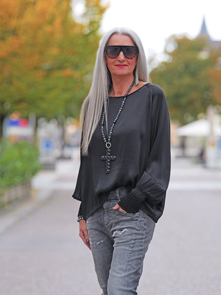 Mature Frau in OFF#DLY LS Shirt Valentina black mit weiten Ärmeln und elegantem Glanz, kombiniert mit grauen Jeans. Accessoires: lange Kette mit Kreuzanhänger, Sonnenbrille. Urbaner Hintergrund.