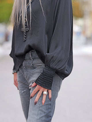 OFF#DLY LS Shirt Valentina anthrazit in fließendem Viskosestoff mit weiten Ärmeln und elastischen Bündchen, kombiniert mit grauen Jeans und auffälligem, kreuzförmigem Perlenanhänger.