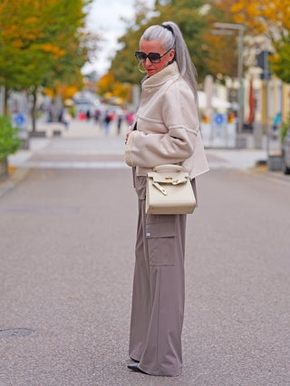 Eine Frau trägt die Penn&Ink Trousers taupe mit weitem Bein und großen Seitentaschen, kombiniert mit einer shearlinggefütterten Jacke und einer cremefarbenen Handtasche.