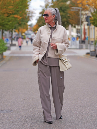 Ältere Frau trägt Penn&Ink Trousers taupe mit weitem Bein und großen Seitentaschen, kombiniert mit einem shearlinggefütterten Jacke und einer strukturierten Ledertasche. Ideal für stilvollen Herbstspaziergang.