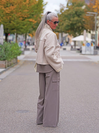 Frau trägt Penn&Ink Trousers taupe mit weitem Schnitt und großen Seitentaschen, während sie in einer städtischen Umgebung mit herbstlichen Bäumen spaziert.