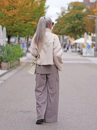 Person von hinten in Penn&Ink Trousers taupe, weit geschnittene Cargohose mit großen Seitentaschen, elastischem Bund, in einer urbanen Herbstlandschaft, kombiniert mit cremefarbener Jacke und strukturiertem Schultertasche.