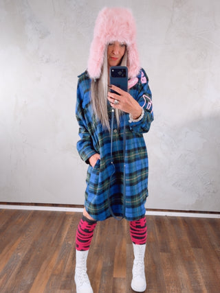 Person in einem langen Hemdkleid mit #VDR Leg Warmers antra/pink, die bis zum Knie reichen, kombiniert mit weißen, geschnürten High-Top-Stiefeln in einem Studio. Das Smartphone verdeckt teilweise das Gesicht.