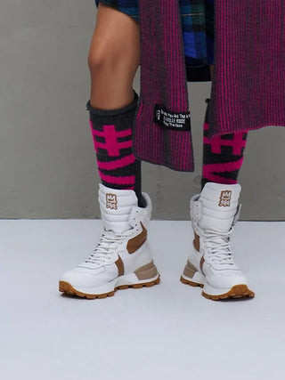 Die #VDR Leg Warmers antra/pink zeigen sich in einem auffälligen schwarz-pink geometrischen Muster, getragen zu kniehohen weißen Stiefeln mit braunen Akzenten und einer dunklen karierten Kleidung.