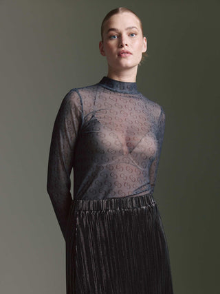 Frau trägt Cotton Candy Floortje II Tee multicolor, ein langärmeliges, transparentes Oberteil mit Kettenmotiv. Sichtbar ist ein hautfarbener Bralette darunter, kombiniert mit einem metallic-schimmernden Faltenrock.