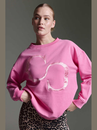 Frau trägt das Cotton Candy. Sweat Lune pink, ein lässiges, superweiches Sweatshirt mit glänzenden C-Formen vorne, kombiniert mit Leopardenmuster-Skinny-Pants. Hände in den Seitenschlitzen des Sweatshirts.