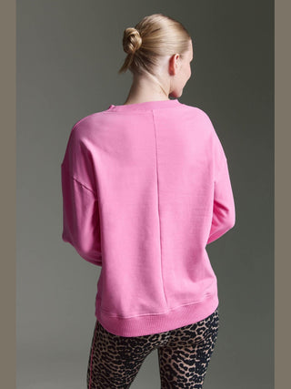 Person von hinten in einem neutralen Hintergrund, trägt das Cotton Candy Sweat Lune pink Sweatshirt mit Rückennaht und Seitenschlitzen, kombiniert mit hochgeschnittenen, leopardengemusterten Leggings. Haare zu einem niedrigen Knoten gebunden.