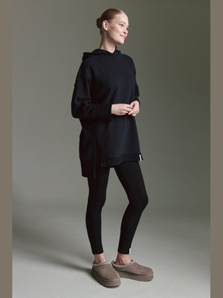 Das Model trägt das Cotton Candy Sweat Luna black, ein oversized Hoodie mit Seitenschlitzen, kombiniert mit schwarzen Leggings und hellbraunen Slip-On-Schuhen. Perfekt für einen lässigen, trendigen Look.