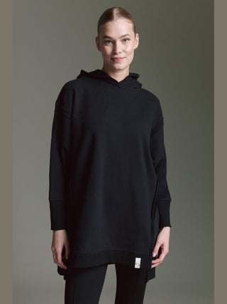 Junge Frau trägt das Cotton Candy Sweat Luna black, ein oversized schwarzes Kapuzen-Sweatshirt mit Seitenschlitzen, kombiniert mit schwarzen Leggings, stehend vor neutralem Hintergrund, lässig und modern.
