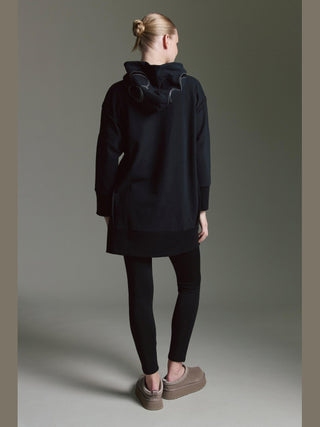 Person von hinten in Cotton Candy Sweat Luna black: langes, schwarzes Hoodie-Kleid mit Kapuze und Seitenschlitzen, kombiniert mit schwarzen Leggings und taupefarbenen Plüschslippern. Haare im ordentlichen Dutt.