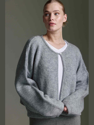 Die Cotton Candy Ona Cardigan grey/mel zeigt eine locker sitzende, hellgraue Strickjacke mit überschnittenen Schultern, langen Ärmeln und einem Knopf am Halsausschnitt, getragen über einem weißen T-Shirt.