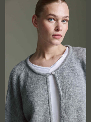 Nahaufnahme einer jungen Frau, die den Cotton Candy Ona Cardigan grey/mel trägt, mit einer Knopfleiste und überschnittener Schulter, über einem weißen Oberteil.
