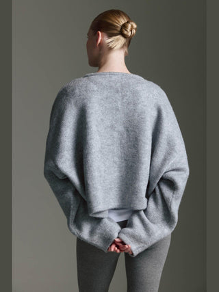 Eine Person trägt den Cotton Candy Ona Cardigan grey/mel mit Knopfleiste und überschnittener Schulter, über einem weißen Oberteil, kombiniert mit grauen Leggings. Haare sind zu einem ordentlichen Dutt gebunden.