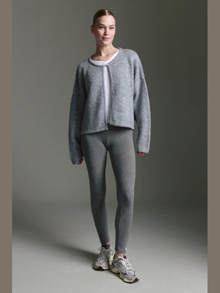 Person trägt den Cotton Candy Ona Cardigan grey/mel, einen weichen, leicht übergroßen Strickcardigan mit Knopfleiste und überschnittener Schulter, kombiniert mit weißen Oberteil und grauen Leggings.