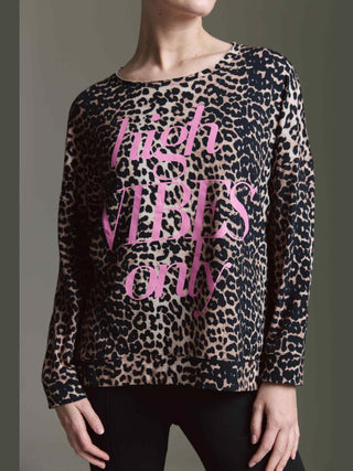 Person trägt das Cotton Candy Tee Irma multicolor, ein langärmeliges, leopardengemustertes Oberteil mit pinkem „high VIBES only“-Schriftzug. Hände ruhen seitlich, Hintergrund schlicht grau.