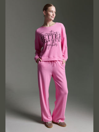 Person trägt Cotton Candy Tee Irma pink mel, ein langärmeliges Oberteil mit Aufdruck, kombiniert mit passenden Loungehosen. Hände in Taschen, Haare zurückgebunden, braune Slipper ergänzen den Look.
