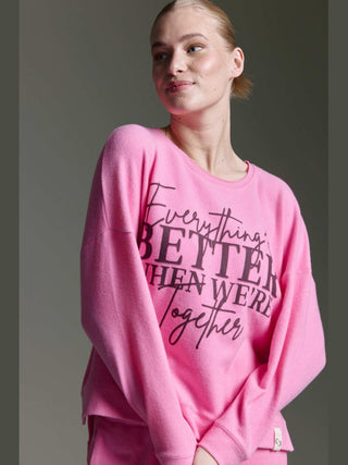 Frau in rosa Cotton Candy Tee Irma pink mel, langärmelig mit dem Aufdruck Everything BETTER when we're Together, steht seitlich, Haare zurückgebunden, vor neutralem Hintergrund.