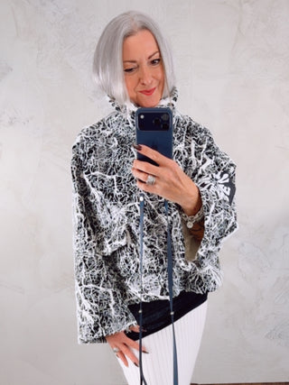 #VDR Sweater 18236 black/white mit Lack-Glanzoberfläche, abstraktem Schwarz-Weiß-Splatter-Print und Stehkragen, getragen von einer Person mit silbergrauem Bob, kombiniert mit weißen, eng anliegenden Hosen.