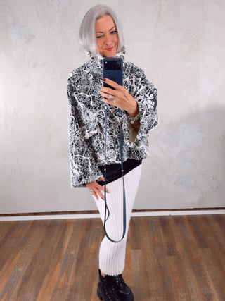 Person im Spiegelselfie trägt den #VDR Sweater 18236 black/white mit Lack-Glanzoberfläche und abstraktem Muster, kombiniert mit weißen, gerippten Leggings und klobigen schwarzen Stiefeln. Minimalistischer Raumhintergrund.