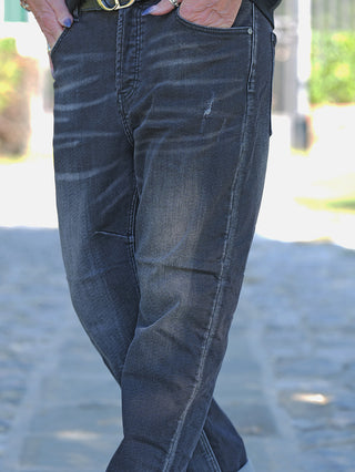 Person trägt OFF#DLY Denim Mira Black Wash Jeans mit leicht abgenutztem Look an Oberschenkeln und Knien, klassischem 5-Pocket-Style und Gürtel mit metallener Schnalle auf steinernem Weg.