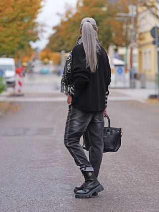Eine Person trägt den Cotton Candy Cardigan Kristi black, ein oversized Strickcardigan mit Knopfleiste und besonderen Ärmelabschlüssen, über einem gestreiften Longsleeve in einer herbstlichen Straßenszene.