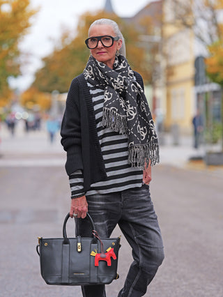 Ältere Frau trägt Cotton Candy Cardigan Kristi black, oversized mit Knopfleiste und besonderem Ärmelabschluss, kombiniert mit dunklen Jeans und einem strukturierten schwarzen Ledertasche in einer herbstlichen Stadtlandschaft.