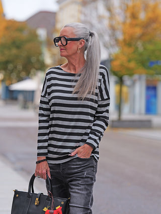 Ältere Frau trägt das OFF#DLY Ringel Dora black/grey Shirt mit rundem Ausschnitt und geschnittenen Kanten, kombiniert mit schwarzen Jeans und einer schwarzen Ledertasche in der Hand.