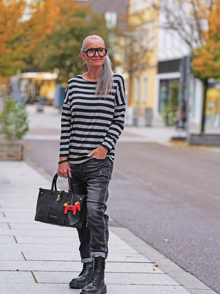 Ältere Frau trägt das OFF#DLY Ringel Dora black/grey mit Rundhals und geschnittenen Kanten, kombiniert mit dunklen Jeans und schwarzen Stiefeln, während sie eine strukturierte schwarze Ledertasche hält.