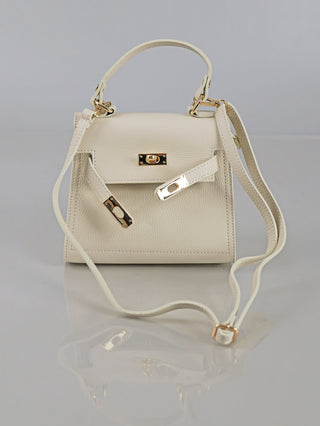 Kleine, strukturierte OFF#DLY Leder Bag creme mit goldfarbenem Drehverschluss, dekorativen Riemenakzenten und verstellbarem Schulterriemen, auf reflektierender Oberfläche präsentiert.