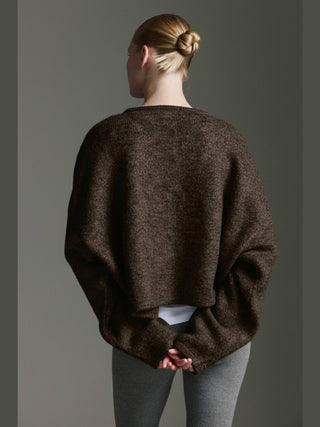 Eine Person trägt den Cotton Candy Ona Cardigan espresso in lockerem, dunklem Strick mit voluminösen Ärmeln, über einem weißen Oberteil, kombiniert mit grauen Leggings, Haare im Dutt gebunden.