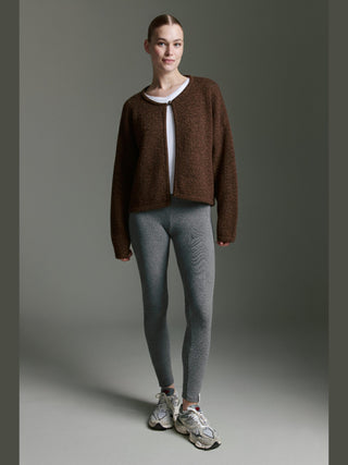 Model trägt den Cotton Candy Ona Cardigan espresso, eine langärmlige, offene Strickjacke mit Knopf. Kombiniert mit weißem Shirt, grauen Leggings und Sneakers, präsentiert sie einen lässig-modernen Look.