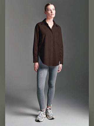 Frau trägt die Cotton Candy Bluse Olivia espresso, eine oversized, langärmelige, dunkelbraune Bluse mit Knopfleiste am Rücken, kombiniert mit grauen Leggings und weißen Chunky-Sneakern, vor neutralem Hintergrund.