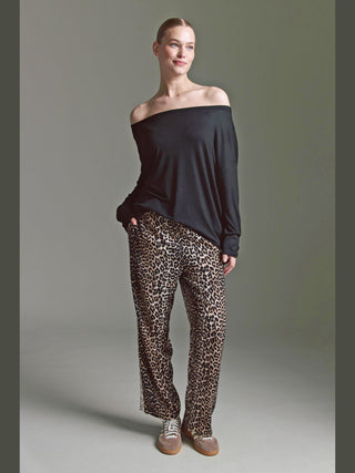 Person trägt Cotton Candy Ondra Tee Black, ein off-shoulder, langärmeliges schwarzes Oberteil, das die Schultern betont. Kombiniert mit lockeren Leopardenhosen und beigen Sneakers, wirkt der Look entspannt und elegant.