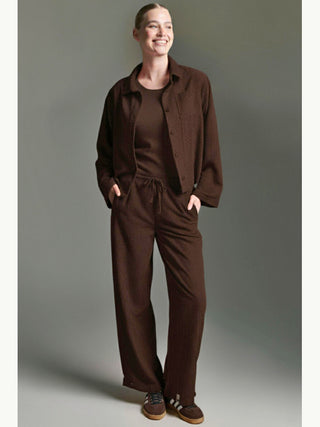 Frau trägt Cotton Candy Bluse Jacke Mesude espresso, locker geschnitten aus weichem, geripptem Cord, über passendem Top und Hose. Hände in Taschen, kombiniert mit braunen Sneakers, vermittelt lässigen Komfort.