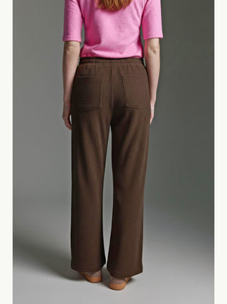 Person von hinten in Cotton Candy Pant Mehtap espresso, einer locker geschnittenen, dunkelbraunen Cordhose mit breiten Beinen und Sitztaschen, kombiniert mit pinkem Oberteil und flachen Mules, entspannter Look.