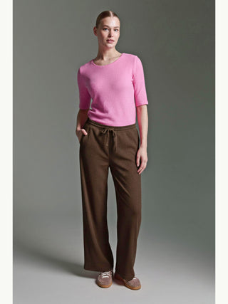 Frau trägt Cotton Candy Pant Mehtap espresso, weiche Cordhose mit weitem Schnitt und elastischem Bund. Kombiniert mit einem rosa Oberteil und braunen, flauschigen Schnürschuhen vor schlichtem Hintergrund.