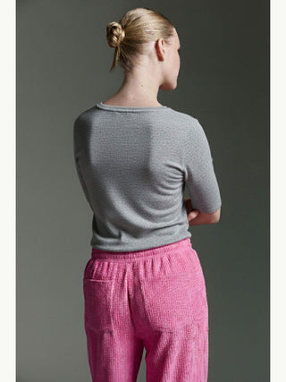 Person von hinten in einem Cotton Candy Tee Bale grey/mel mit halblangen Ärmeln, kombiniert mit lockeren, texturierten rosa Hosen. Haare im Dutt, Arme lässig am Rücken verschränkt.