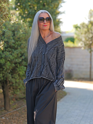 Ältere Frau trägt die OFF#DLY West Ada black wash Strickweste mit markantem Strickmuster und oversized Look, kombiniert mit weiten schwarzen Hosen. Die Weste fällt locker und modern über die Schulter.