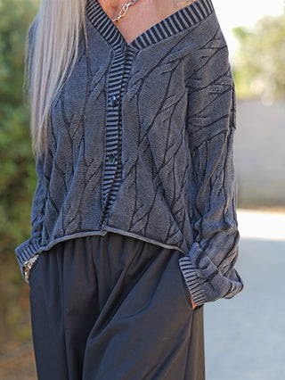 Person trägt eine OFF#DLY West Ada black wash Strickweste mit markantem Strickmuster, V-Ausschnitt, Knopfleiste und goldener Kette, kombiniert mit lockeren schwarzen Hosen, zeigt modernen, lässigen Stil.