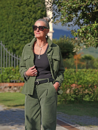 Die reife Frau trägt das OFF#DLY Cord Hemd Violetta grün mit offenen Saumkanten, kombiniert mit einer schwarzen Hose. Ihr eleganter Look wird durch Accessoires wie Sonnenbrille und Schmuck ergänzt.