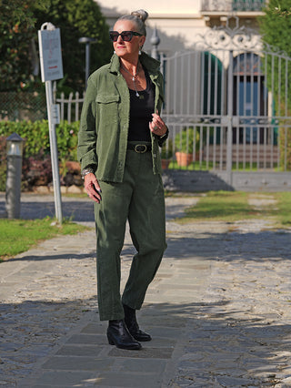 Frau trägt das OFF#DLY Cord Hemd Violetta in Olive, offen über einem schwarzen Tanktop, kombiniert mit passender Hose, zeigt lässigen Chic vor einer Villa mit Tor, ideal für modische Alltagslooks.