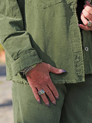Nahaufnahme des OFF#DLY Cord Hemd Violetta grün mit offenen Saumkanten, getragen mit passenden Cordhosen. Eine Hand mit ombré-Nägeln und silbernem Ring, die andere justiert das Hemd.