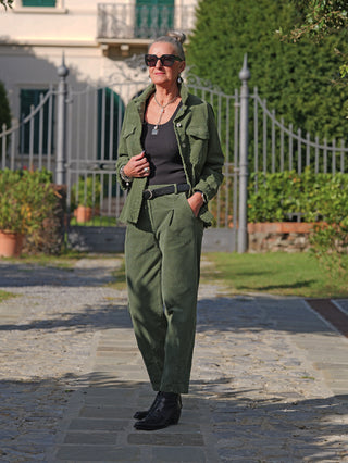 Ältere Frau trägt die OFF#DLY Cord Pant Veronica grün, kombiniert mit einem schwarzen Tanktop und olivegrüner Cordjacke, vor einer dekorativen Toranlage, betont durch schwarze Accessoires und silbernen Schmuck.