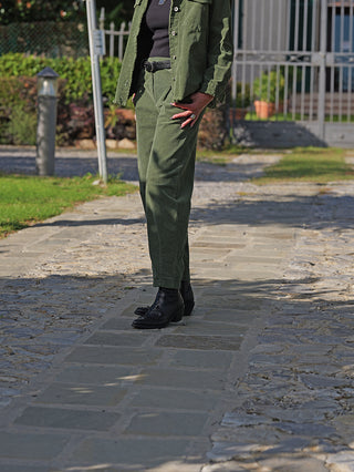 Person trägt OFF#DLY Cord Pant Veronica grün, ein leicht texturiertes, hochtailliertes Outfit, kombiniert mit schwarzem Gürtel und Stiefeln. Die Hand in der Tasche verleiht einen lässigen Look.