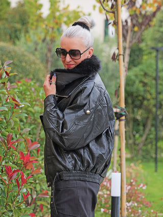 Frau in Garten trägt die OFF#DLY Jacke Bria black, eine glänzende Bomberjacke mit abnehmbarem Kunstfellkragen, oversized-Schnitt und goldenen Details, hält den Kragen mit Ringen geschmückten Fingern.