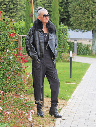 Ältere Frau trägt die OFF#DLY Jacke Bria black mit Fellkragen und goldenen Details, kombiniert mit einer schwarzen Hose und Stiefeln, auf einem Gartenweg stehend, Hände in den Taschen.