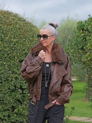 Ältere Frau im Garten trägt die OFF#DLY Jacke Bria braun, eine oversized Kunstlederjacke mit Fellkragen und goldfarbenen Details, kombiniert mit einem schwarzen Top und dunklen Hosen.