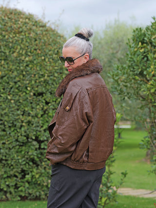 Eine Person trägt die OFF#DLY Jacke Bria braun, eine modische Pilotenjacke mit Kunstfellkragen und goldenen Details, in einem Park. Die Jacke verleiht dem Look eine stilvolle, urbane Note.